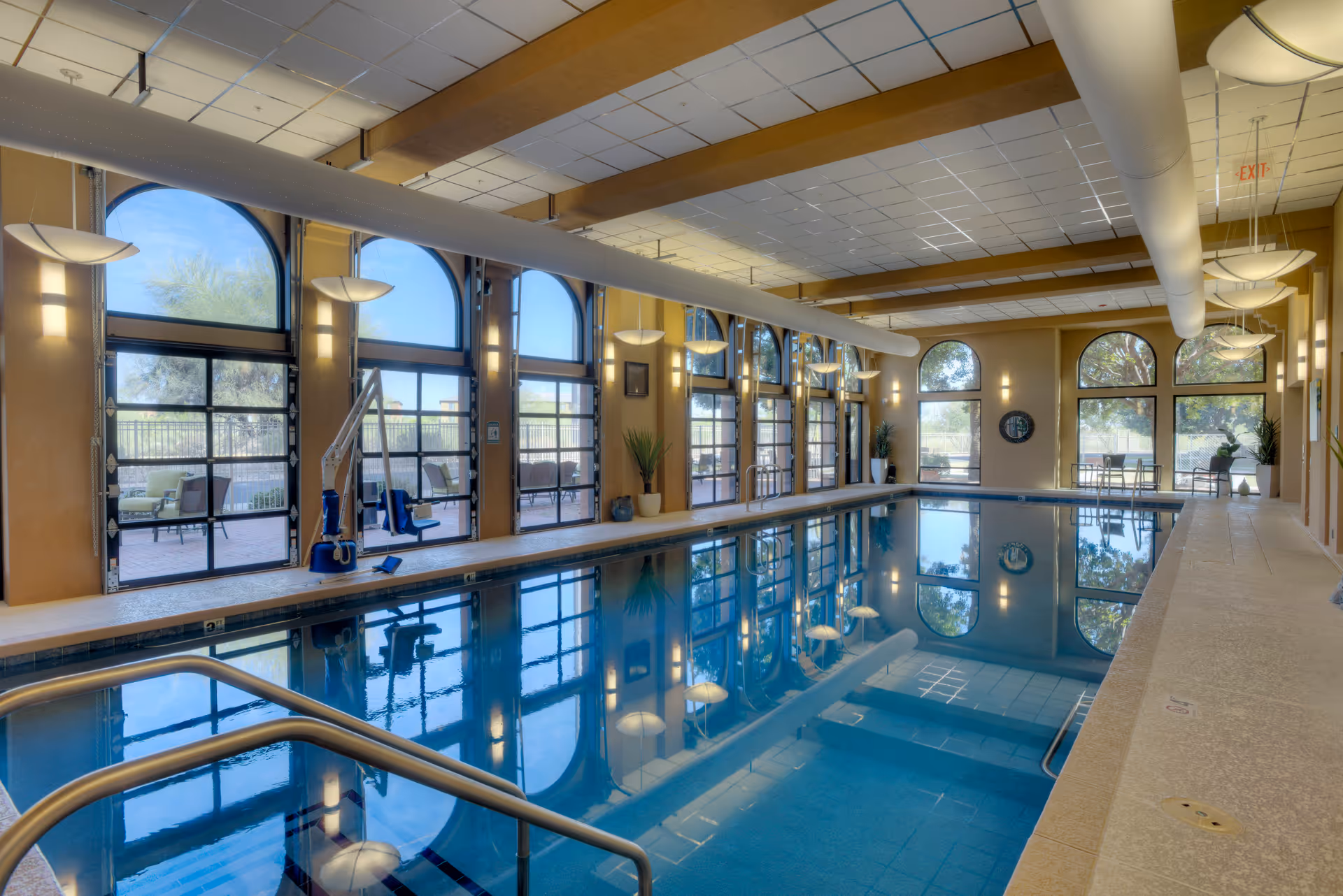 Indoor swimming pool area with large arched windows letting in natural light. The pool is calm and reflects the ceiling lights and windows. There are poolside chairs and plants along the walls, with a pool lift for accessibility.
