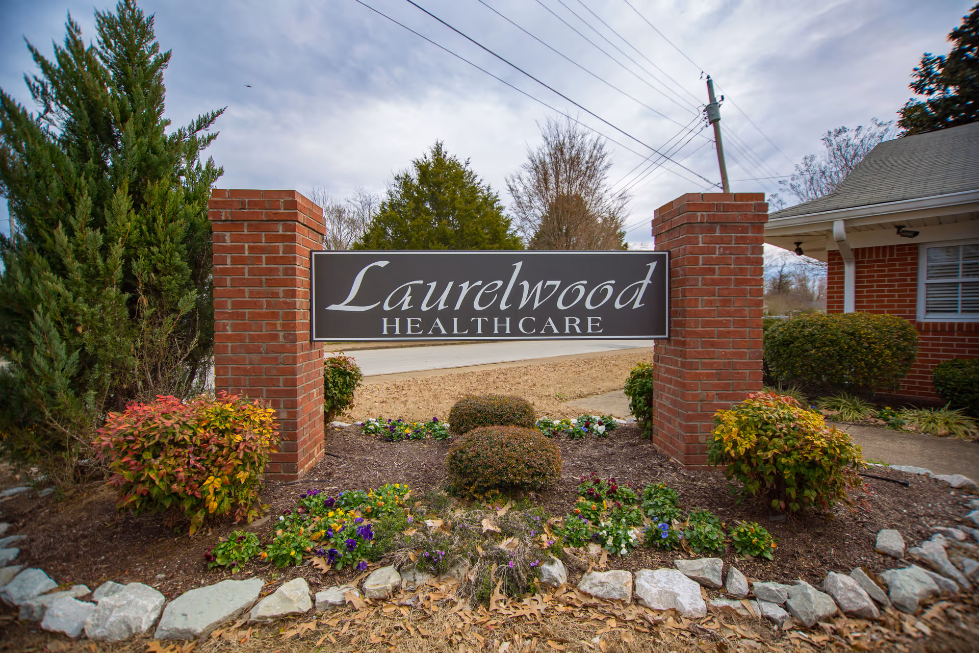 A brick signpost with a black and white sign reading 'Laurelwood Health Care' surrounded by landscaped bushes and flowers, with a building and trees in the background under a cloudy sky.