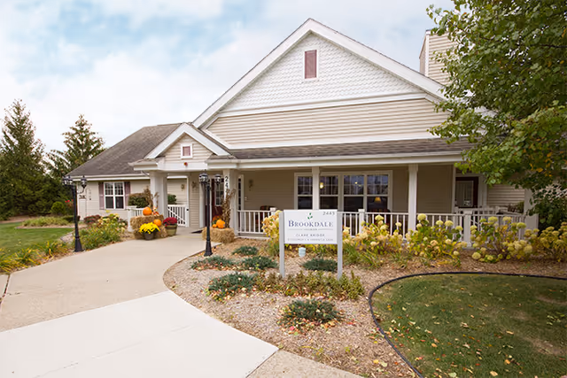 Exterior view of a senior living facility building with a beige facade, white trim, and a covered porch. There is a paved walkway leading to the entrance, landscaped garden beds with plants and flowers, and a sign in front that reads 'Brookdale Clare Bridge'. The scene includes a lamppost and some autumn decorations like pumpkins.
