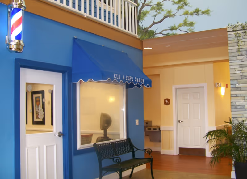 Indoor hallway area of a senior living facility featuring a blue wall with a door and window under a blue awning labeled 'Cut & Curl Salon'. A black metal bench is placed in front of the wall. To the right, there is a beige wall with a white door and a water fountain. A potted plant is visible on the far right, and a mural of a tree is painted on the upper wall.