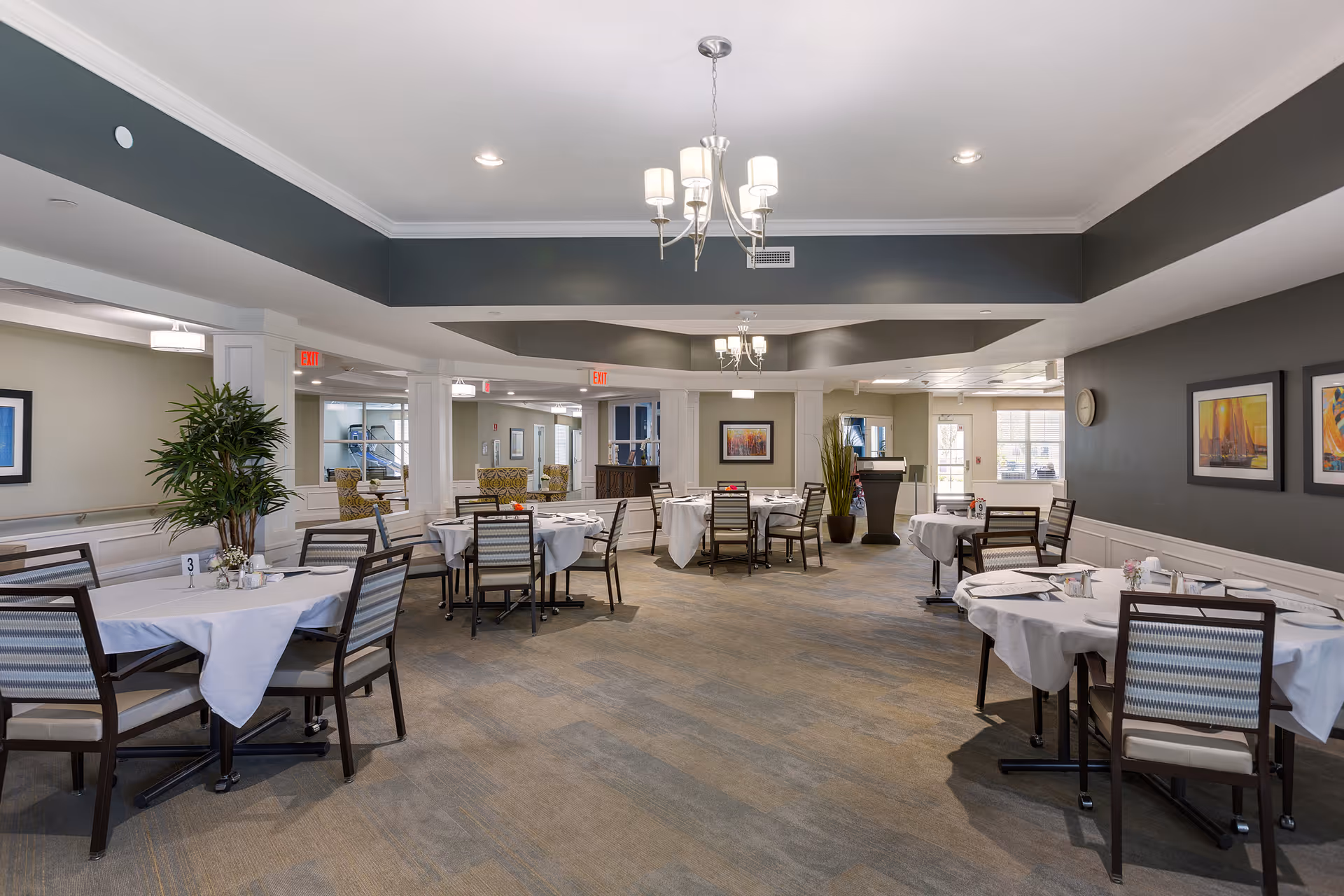 A spacious dining room with multiple round tables covered with white tablecloths, each surrounded by chairs. The room features a modern chandelier, framed artwork on the walls, large windows allowing natural light, and decorative plants. The overall decor is elegant and welcoming.