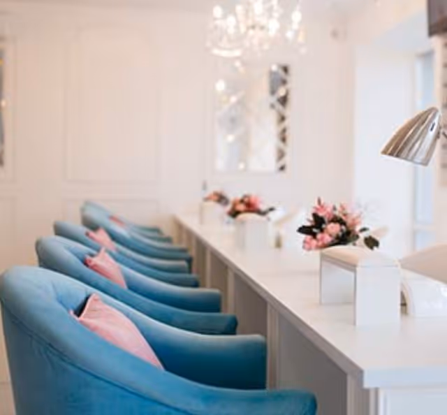 A row of blue upholstered chairs with pink pillows lined up along a white counter table. The table has small floral arrangements and modern desk lamps. The background features a chandelier and a mirror on a white wall.