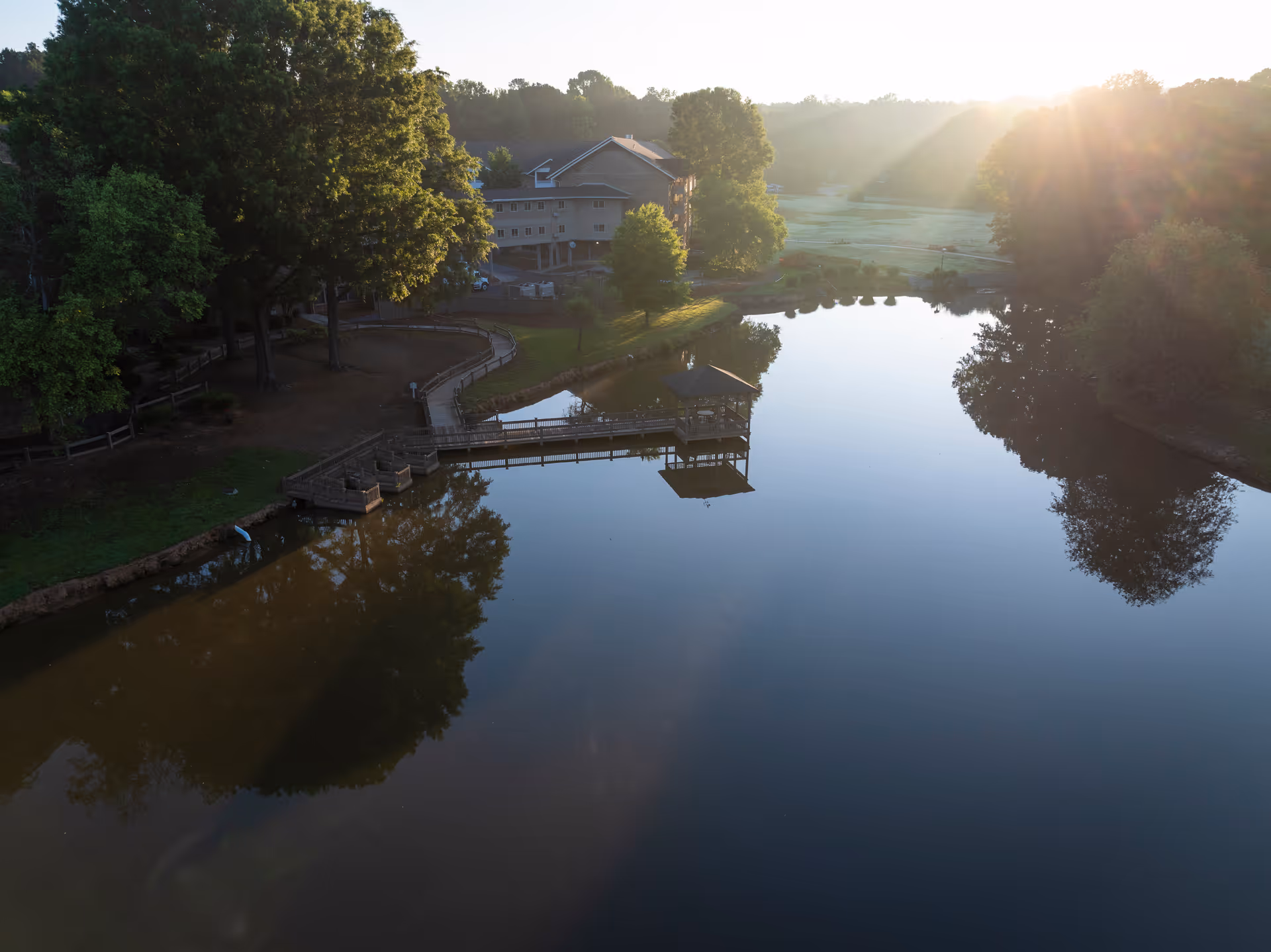 A serene outdoor scene at Presbyterian Village featuring a calm pond with a wooden dock and gazebo extending over the water. Surrounding the pond are large trees and a walking path. In the background, a building is partially visible with sunlight streaming through the trees.