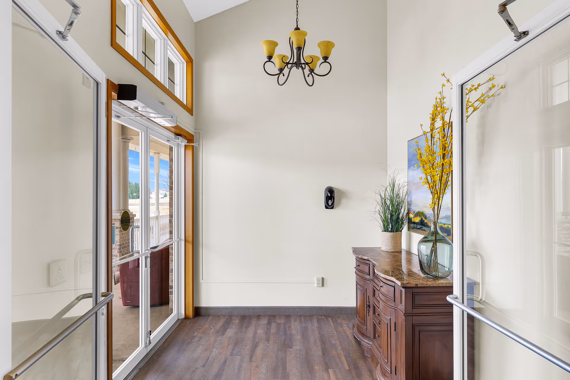 Entrance area of a facility with double glass doors, a wooden sideboard with decorative plants and a painting on the wall, a chandelier hanging from the ceiling, and a view of an outdoor patio with columns visible through the doors.