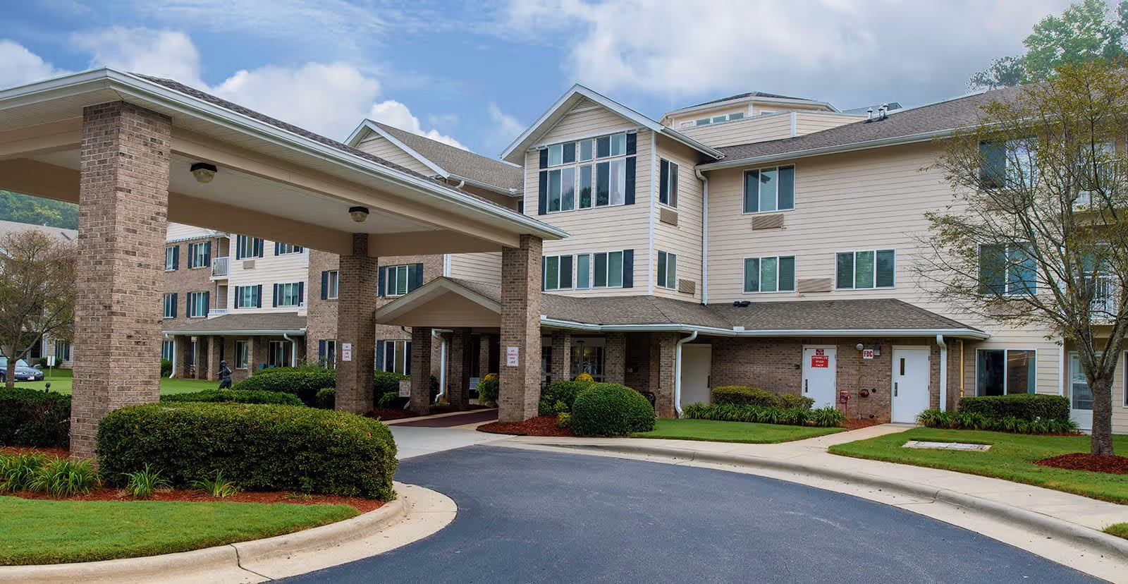 Exterior view of a multi-story senior living facility with beige siding and brick accents. The building has multiple windows and a covered entrance driveway with columns. There are well-maintained shrubs, grass, and trees around the building under a partly cloudy sky.