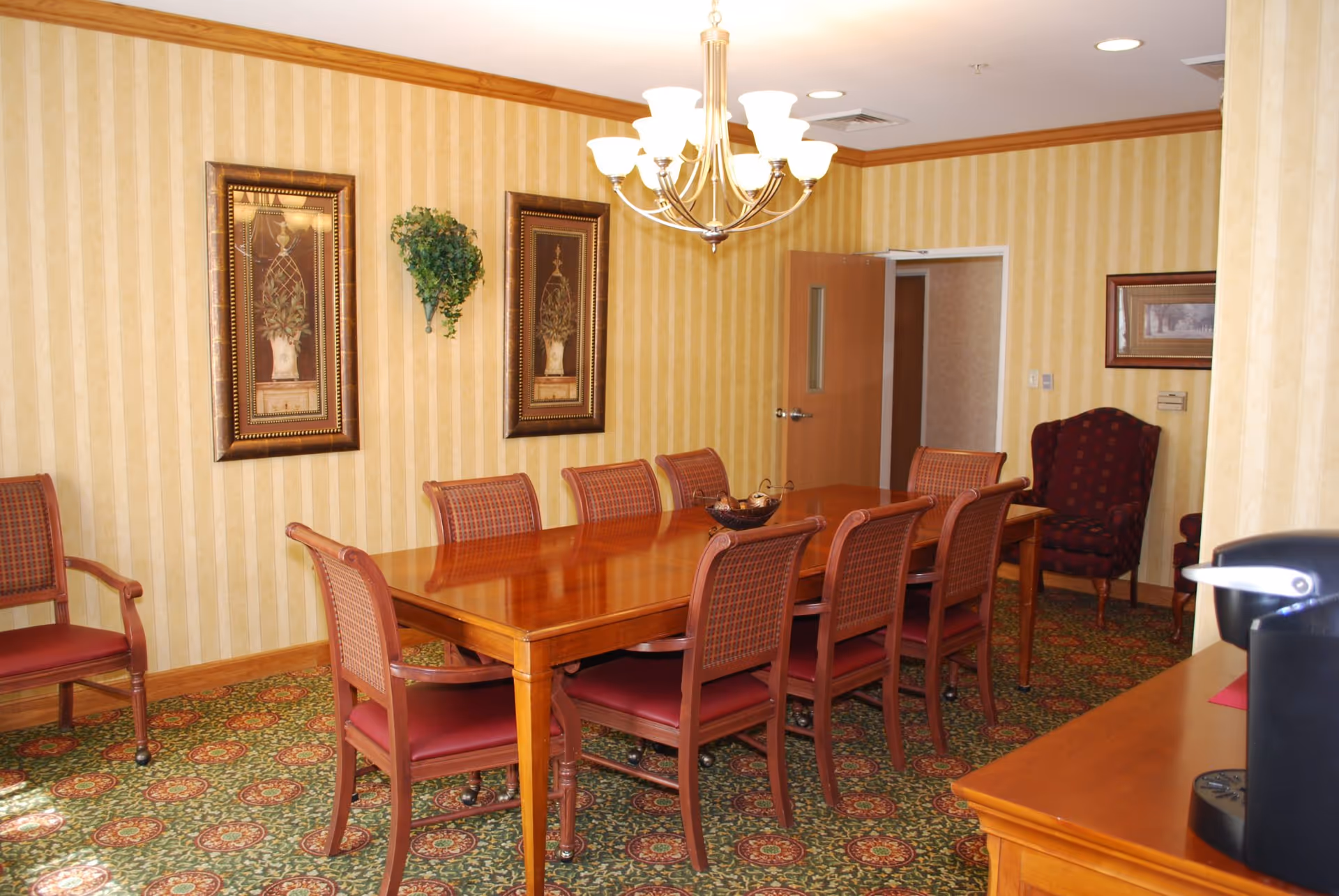 A formal dining room with a long wooden table surrounded by eight chairs with red cushions. The room has patterned carpet, striped yellow wallpaper, two framed botanical prints, a wall-mounted plant, a chandelier with multiple lights, and a wooden sideboard with a coffee maker on it. There is also a burgundy upholstered armchair in the corner and an open door leading to another room.