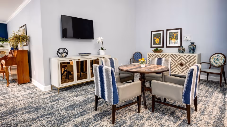 A cozy living room area with a round wooden table surrounded by four upholstered chairs with blue and gray striped backs. A white sideboard with mirrored doors is against the wall beneath a mounted flat-screen TV. Two framed floral artworks and decorative vases are on another sideboard in the background. The room has patterned carpet flooring and a piano is visible on the left side.