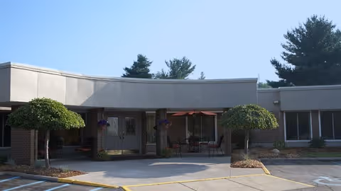 Exterior view of Medilodge of Kalamazoo building entrance with a covered drop-off area, two trimmed trees, and outdoor seating with an umbrella on a clear day.