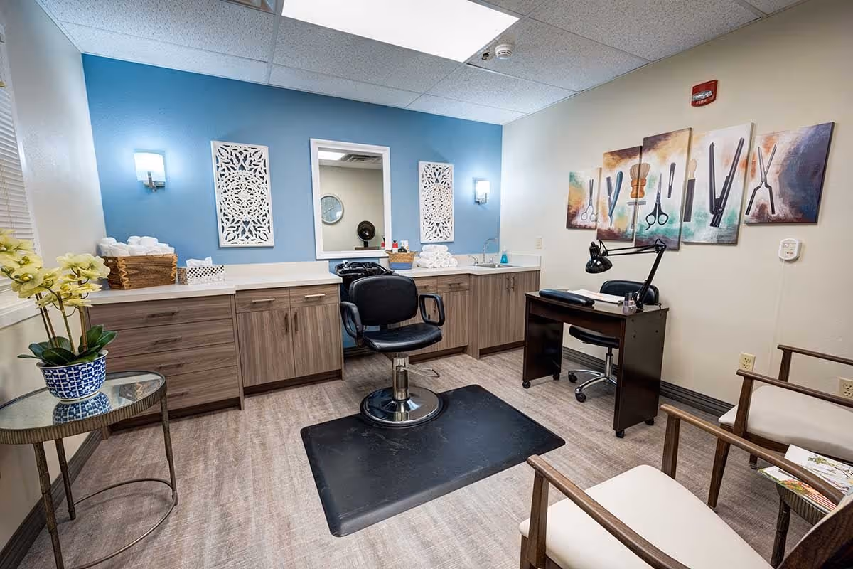 A well-lit salon room with a black styling chair on a black mat in front of a large mirror mounted on a blue accent wall. The room has wooden cabinets with white countertops, a small sink, and neatly folded white towels. There is a small black desk with a lamp and chair, two beige armchairs, a round glass side table with a potted orchid, and colorful wall art depicting hair styling tools.