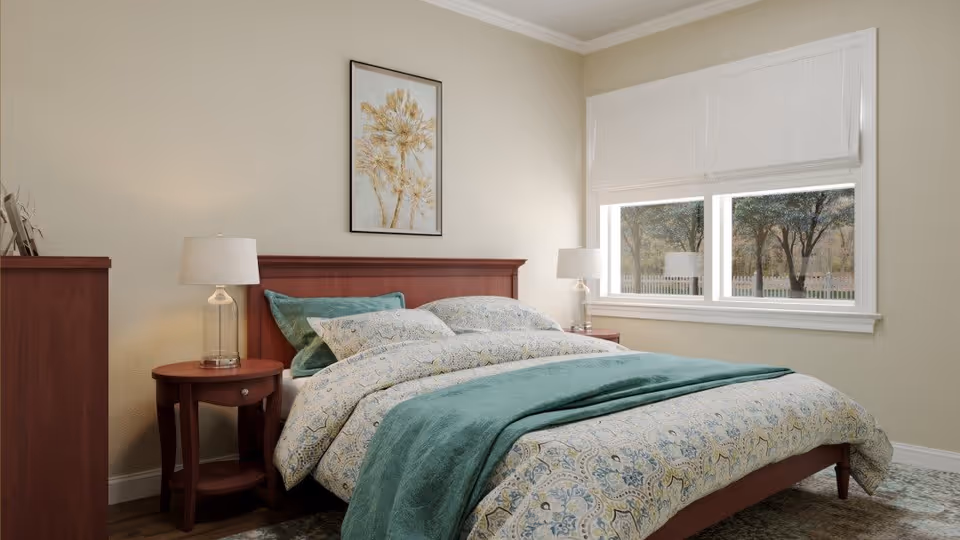 A cozy bedroom with a wooden bed frame, patterned bedding in shades of blue and beige, two matching wooden nightstands each with a lamp, a wooden dresser, a framed botanical artwork on the wall, and a window with white blinds showing trees outside.