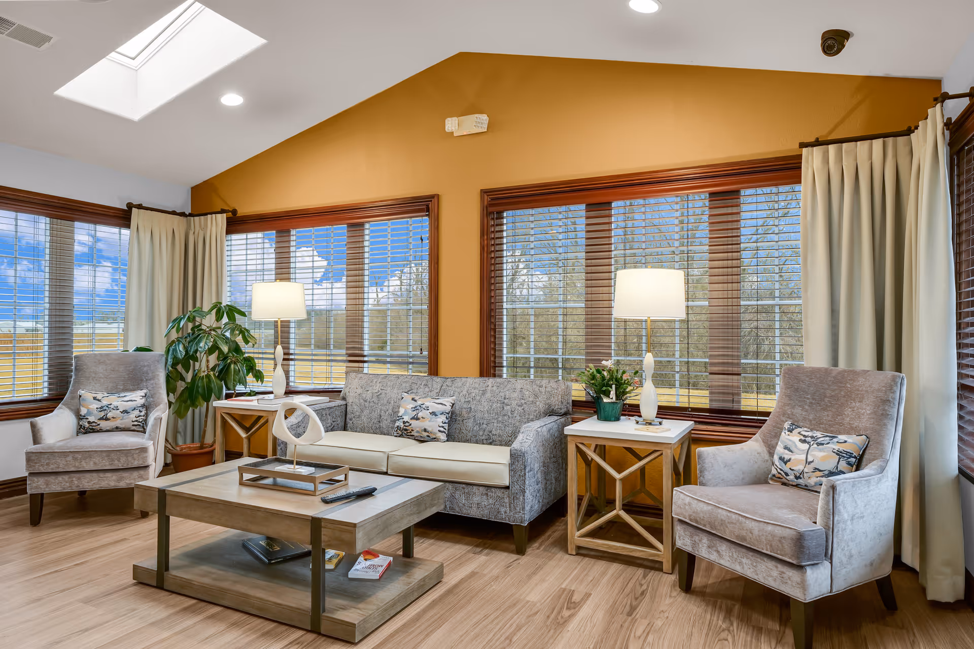 Bright living room with a sofa, two armchairs, coffee table, side tables with lamps, and large windows with blinds.