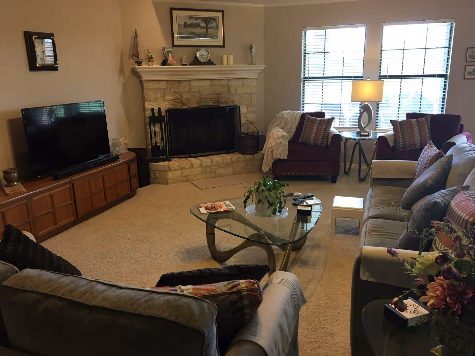 A cozy living room with a stone fireplace in the center, flanked by two purple armchairs with patterned pillows and a side table with a lamp. A glass coffee table with a plant and magazines sits on a beige carpet. A large flat-screen TV is on a wooden cabinet to the left. A gray sofa with pillows is in the foreground, and a window with blinds lets in natural light.