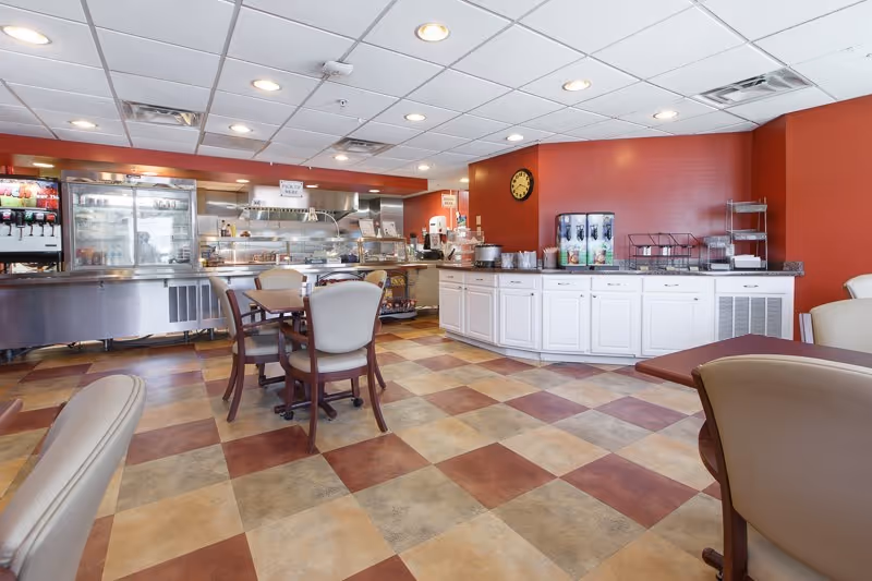 A dining area with several tables and chairs, a beverage dispenser, and a food service counter with a 'Pick Up Here' sign. The room has a checkered tile floor in shades of brown and beige, red walls, and a white ceiling with recessed lighting.