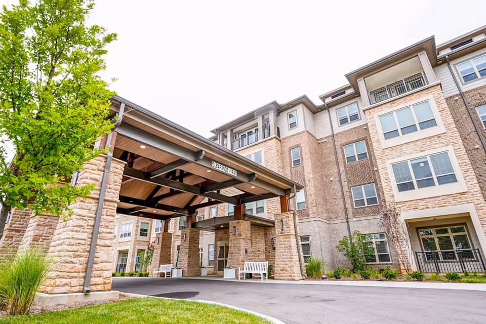 Exterior view of a multi-story senior living facility building with a covered entrance supported by stone pillars. There are benches near the entrance and landscaping with green grass, bushes, and a tree. The sky is overcast.