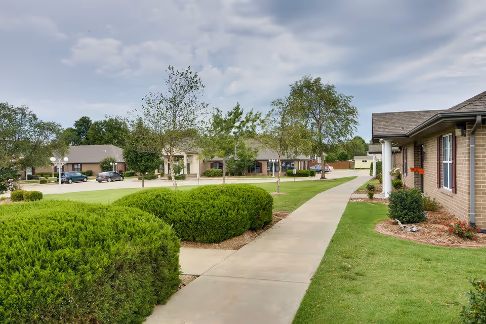 A paved walkway runs through a well-maintained outdoor area of a senior living facility with green bushes, trees, and grass on both sides. Single-story brick buildings with windows and small porches line the path. Several parked cars are visible in the background under a cloudy sky.