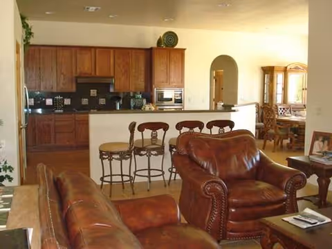 Interior view of a living area with brown leather sofas and armchairs in the foreground, a kitchen with wooden cabinets and a breakfast bar with three high chairs in the background, and a dining area visible through an arched doorway.