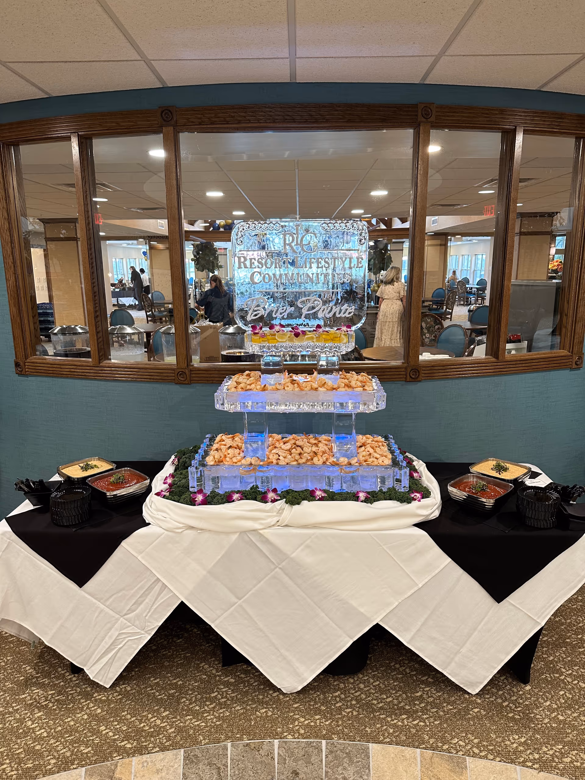 A buffet table with a three-tiered ice sculpture display filled with shrimp, surrounded by bowls of dipping sauces. The ice sculpture has the text 'Resort Lifestyle Communities Brier Pointe' carved into it. The table is covered with white and black tablecloths and decorated with purple flowers. Behind the table is a large window showing a dining area with people and tables.