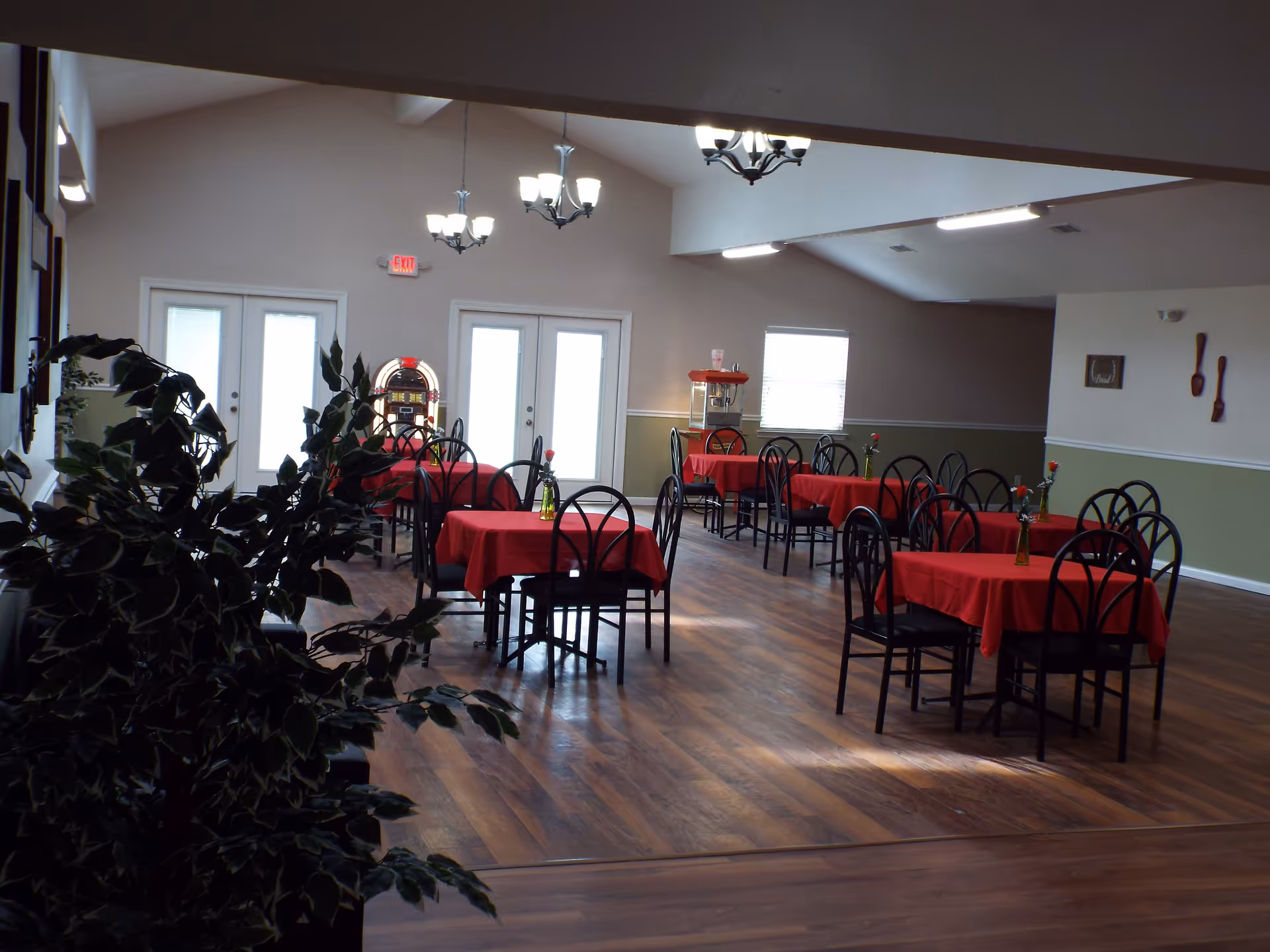 Interior view of a dining room with several tables covered in red tablecloths, each table surrounded by black chairs. Each table has a small vase with a single flower. The room has wooden flooring, light-colored walls with a green lower half, and multiple chandeliers hanging from the ceiling. There are double doors and windows letting in natural light, and a popcorn machine is visible in the background.