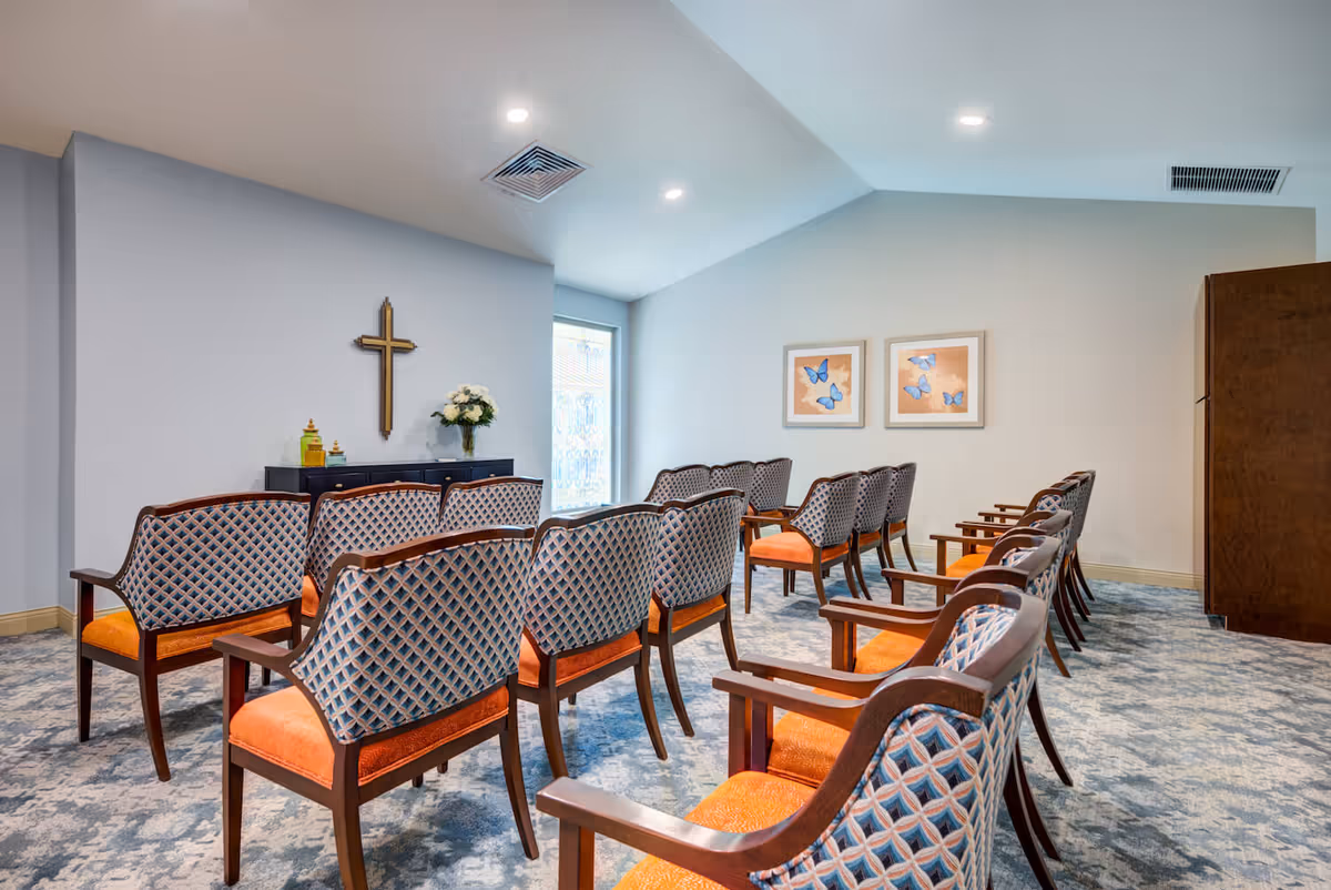A small chapel or meditation room with rows of wooden chairs featuring orange cushions and patterned backs. A wooden cross is mounted on the wall above a black cabinet with decorative items and a vase of white flowers. Two framed pictures of blue butterflies hang on the adjacent wall. The room has a light blue carpet and soft lighting from ceiling fixtures.