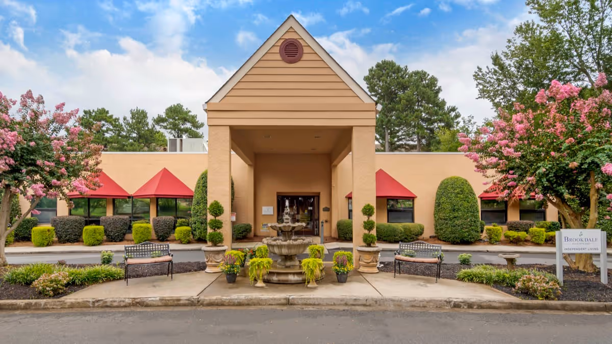 Front exterior view of Brookdale Charlotte East facility with a beige building, red awnings over windows, a central covered entrance, a tiered water fountain, benches, and landscaped greenery including flowering trees and shrubs.