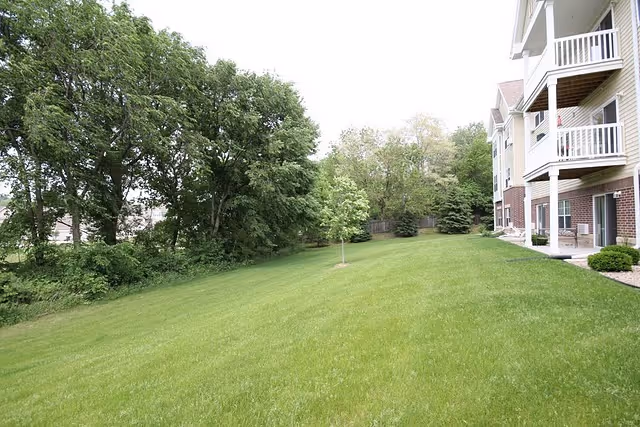 A grassy outdoor area next to a multi-story building with balconies. The area includes a well-maintained lawn, a few small trees, and larger trees in the background.
