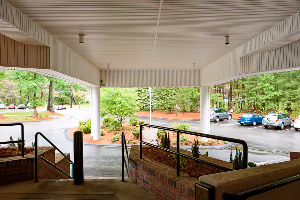 View from under a covered entrance area looking out onto a parking lot with several parked cars and surrounding greenery including trees and shrubs. The entrance has brick walls, metal railings, and a ramp for accessibility.