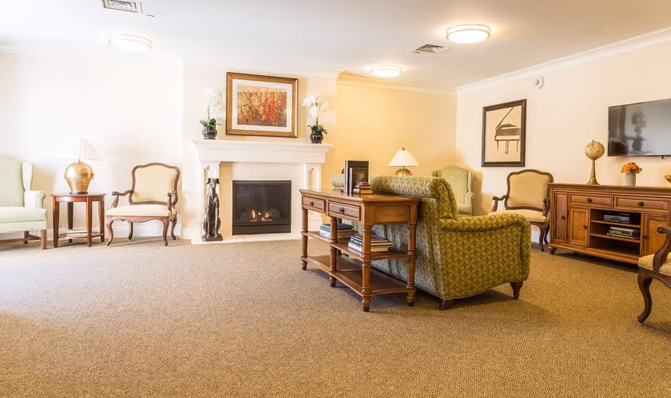 A cozy senior living room with a fireplace, two green upholstered armchairs, two wooden chairs with cushions, a wooden console table with books, a wooden TV stand with a flat-screen TV, decorative lamps, and framed artwork on the walls.