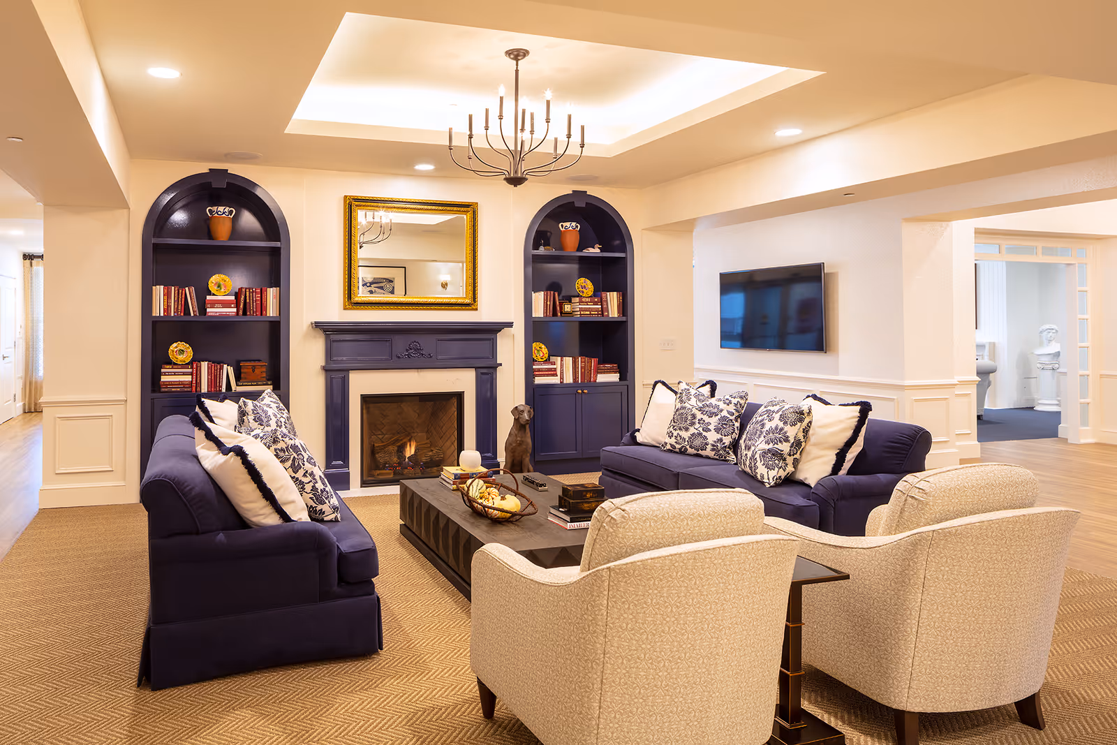 A cozy living room with two navy blue sofas adorned with patterned and plain white pillows, two beige armchairs, a dark wooden coffee table with decorative items, a fireplace with a golden framed mirror above it, and built-in dark blue bookshelves on either side filled with books and decorative plates. The room has warm lighting and a chandelier hanging from a recessed ceiling.