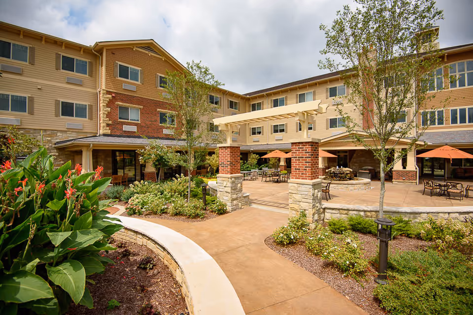 Paved courtyard with seating, umbrellas, landscaping and a fountain in front of a multi-story senior living building.