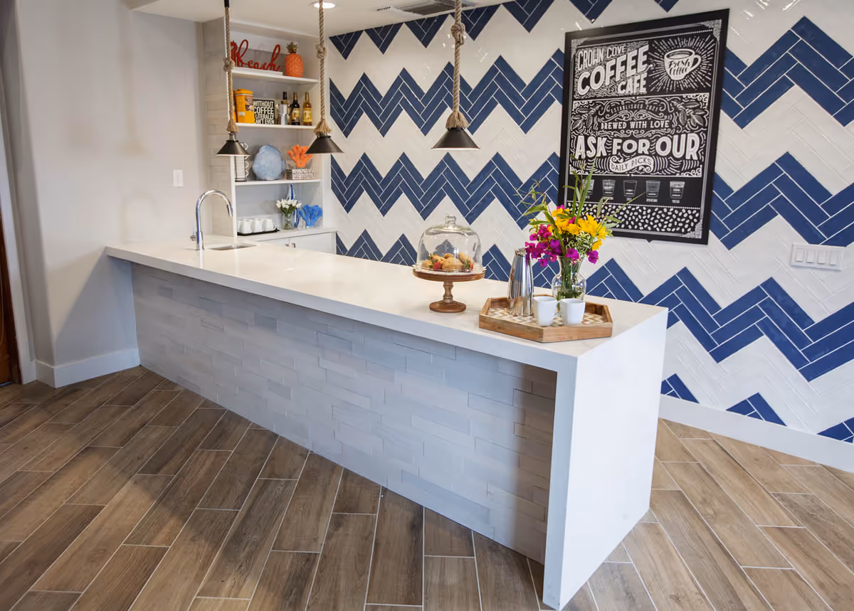 Bright coffee bar with a white island, wood-look floor, blue-and-white chevron tile wall, pendant lights, flowers, and a chalkboard-style 'Crown Cove Coffee Cafe' sign.