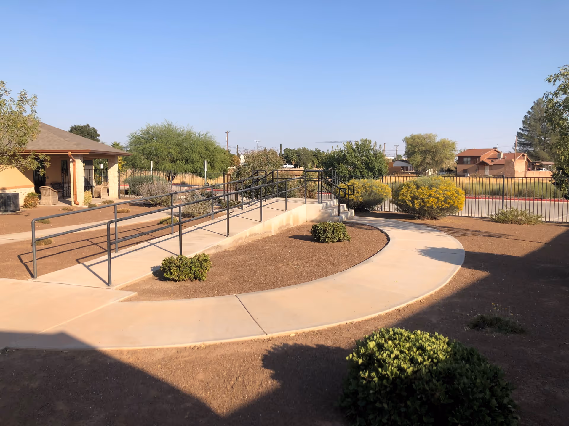 Outdoor view of a senior living facility showing a concrete walkway with a wheelchair ramp and handrails, surrounded by small bushes and trees under a clear blue sky. A building with a covered porch and chairs is visible on the left side.