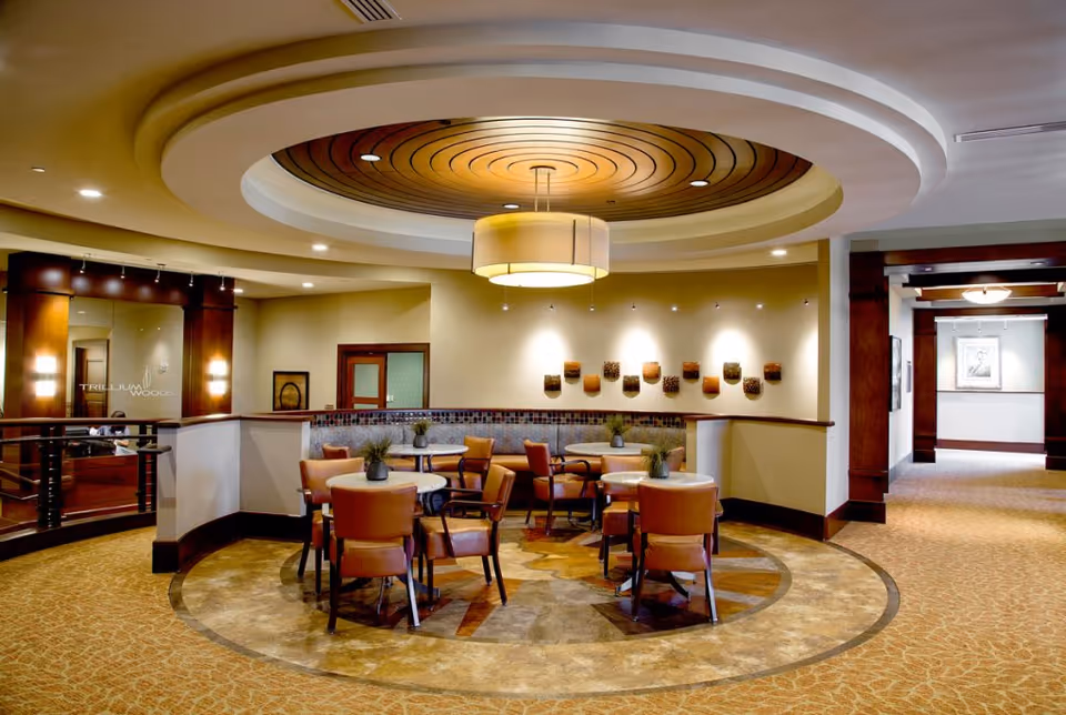 A cozy seating area in a senior living facility with round tables and cushioned chairs arranged on a circular patterned floor. The ceiling features a decorative circular design with a large hanging light fixture. The walls are adorned with small decorative baskets and soft lighting. The space has warm tones with wood accents and carpeted flooring.