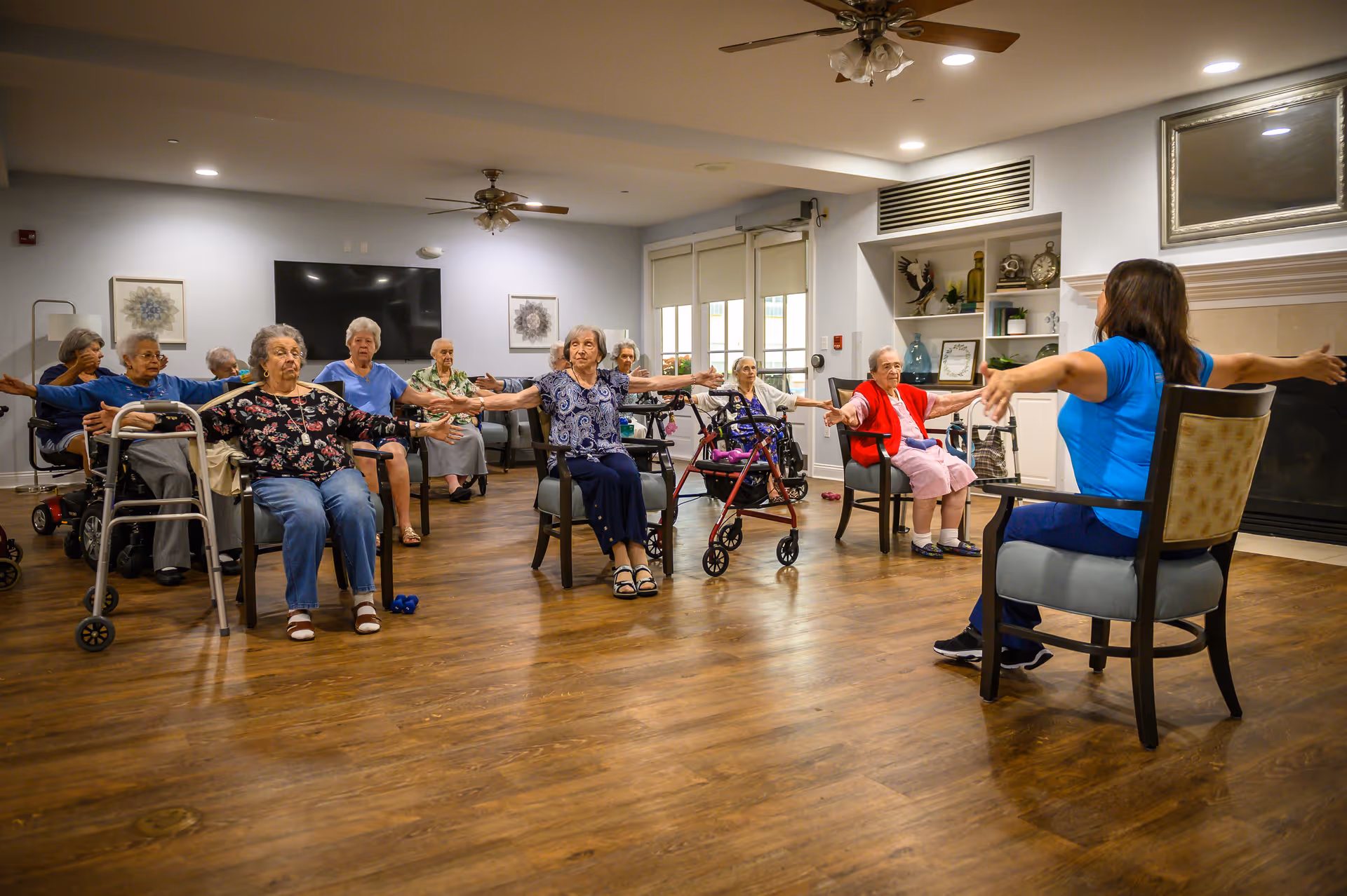 A group of elderly women seated in chairs in a spacious room participating in a seated exercise class led by an instructor. The women have their arms extended to the sides, and some use walkers or wheelchairs. The room has wooden floors, a ceiling fan, a large TV on the wall, and decorative shelves with various items.