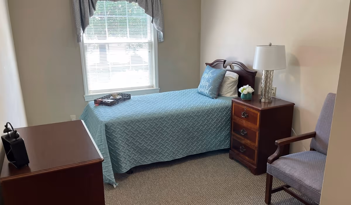 Small single bedroom with a twin bed covered in a light blue quilt, a wooden nightstand with a lamp and flowers, a dresser, and a chair by a window.
