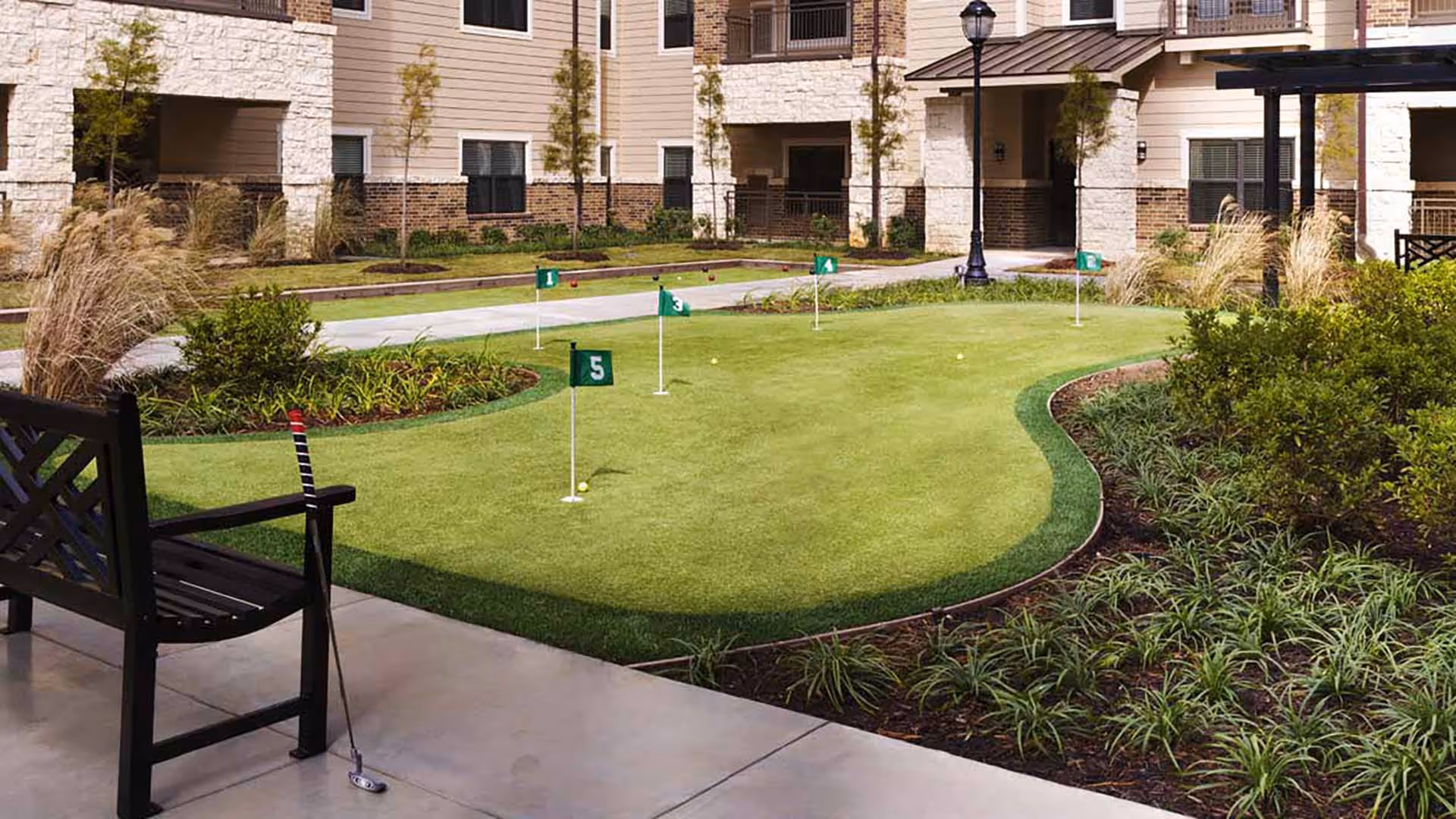 Courtyard putting green with numbered flags, a bench, and landscaped beds in front of a multi-story senior living building.