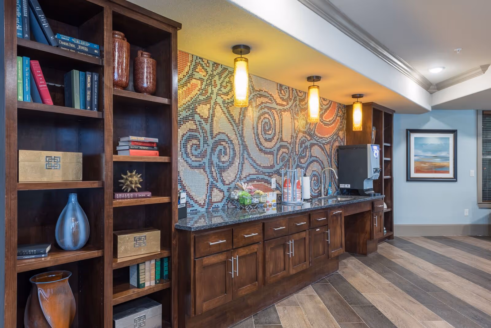 A communal kitchenette with dark wood cabinets and shelves, a mosaic tile backsplash, pendant lights, and a countertop coffee/refreshment station.