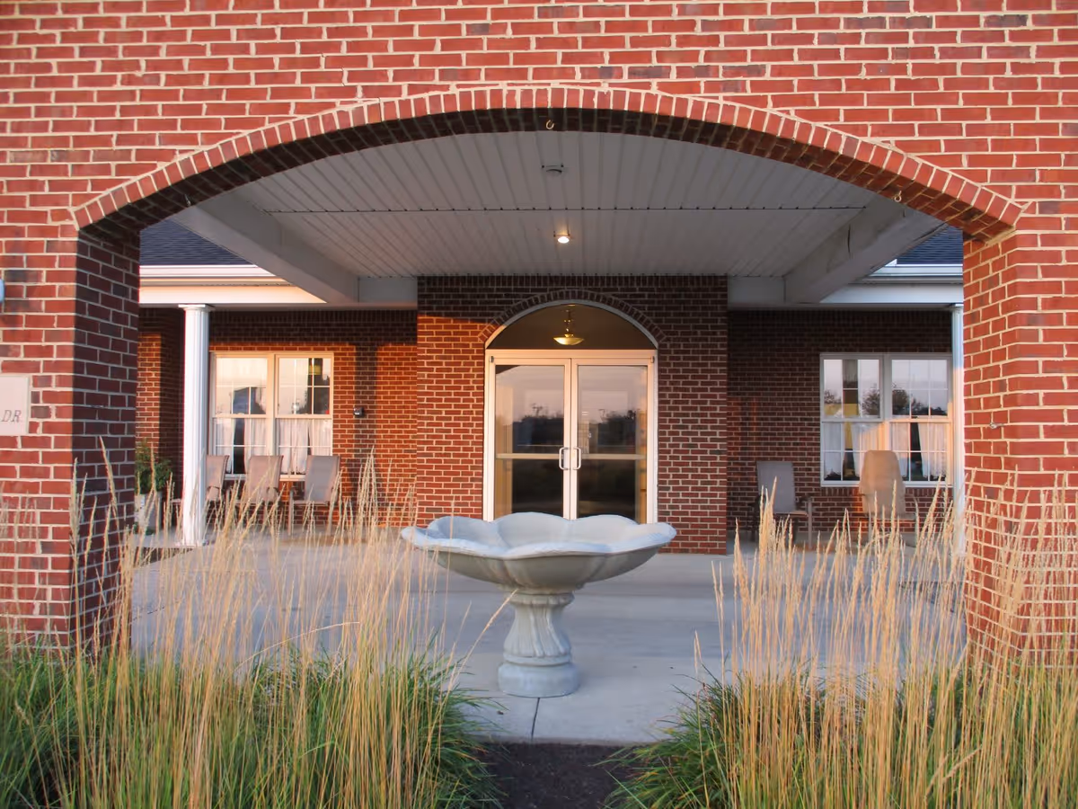 Entrance of a brick building with a covered porch area featuring a large archway. In front of the entrance, there is a decorative stone birdbath surrounded by tall ornamental grasses. The building has large windows with curtains and several chairs placed on the porch.
