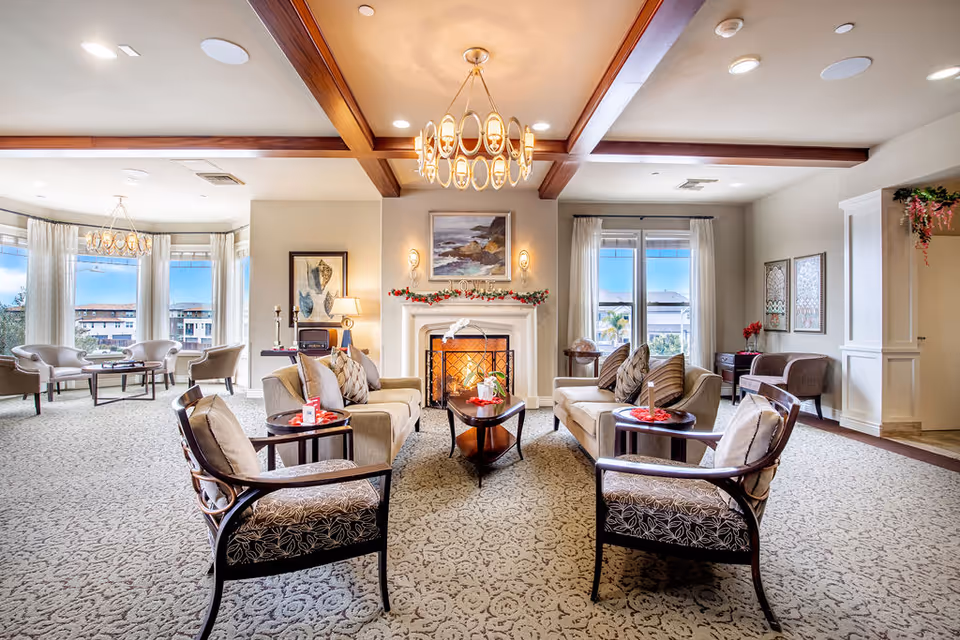 A spacious and well-lit living room area with beige sofas and patterned armchairs arranged around a coffee table. A fireplace decorated with garland and a painting above it is centered on the far wall. Large windows with sheer curtains allow natural light to fill the room. The ceiling features wooden beams and a modern chandelier. Additional seating is visible near the windows on the left side.