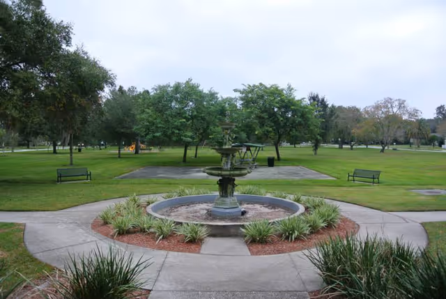 A green outdoor park area with a multi-tiered stone fountain in the center surrounded by plants and a circular paved walkway. There are benches on either side of the walkway and trees in the background under a cloudy sky.