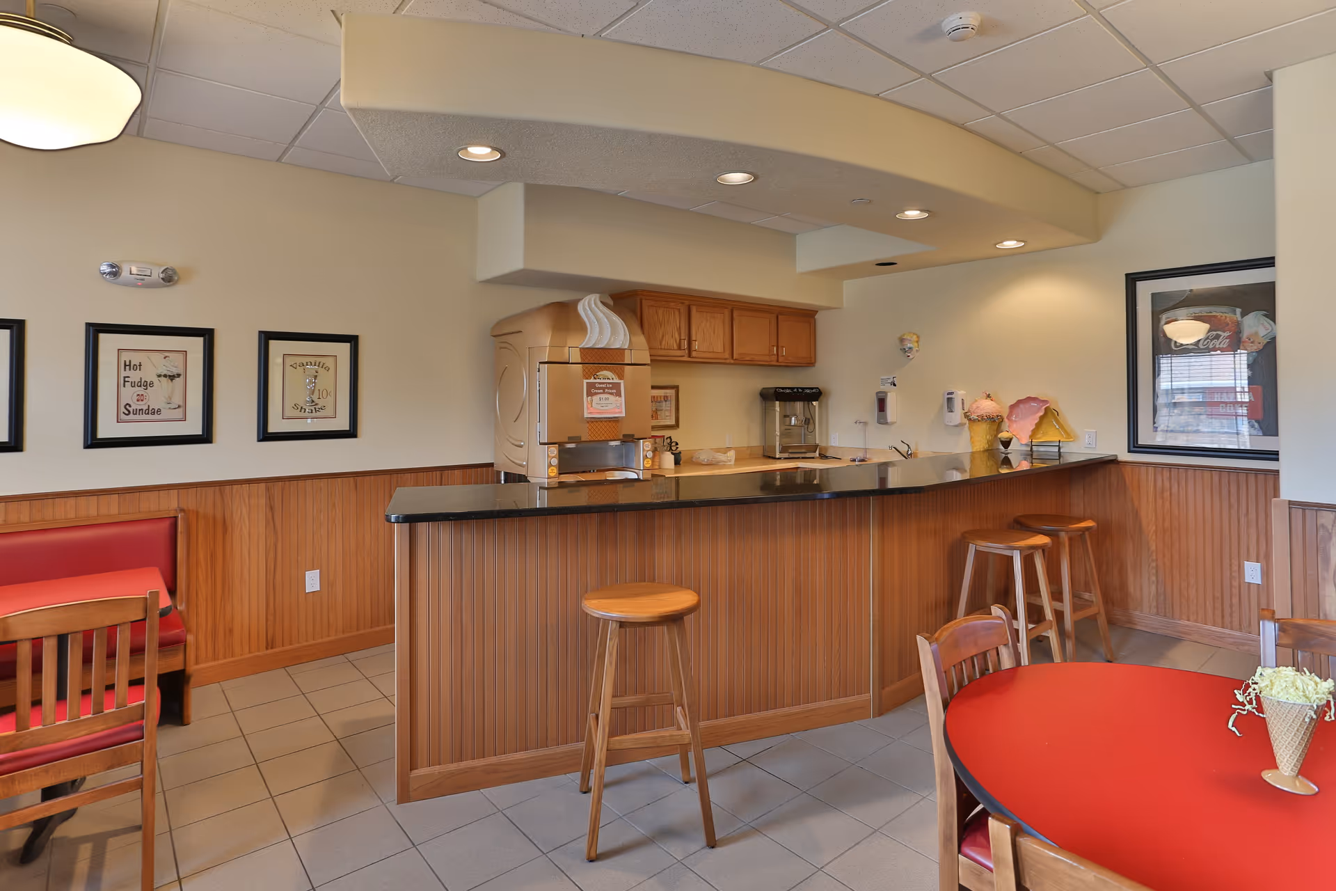 A cozy dining area with a wooden counter and three wooden stools. The counter has a soft-serve ice cream machine and a coffee maker. There are framed vintage-style posters on the walls, a red cushioned bench, wooden chairs, and a round red table with a decorative ice cream cone centerpiece. The room has tiled flooring and a drop ceiling with recessed lighting.