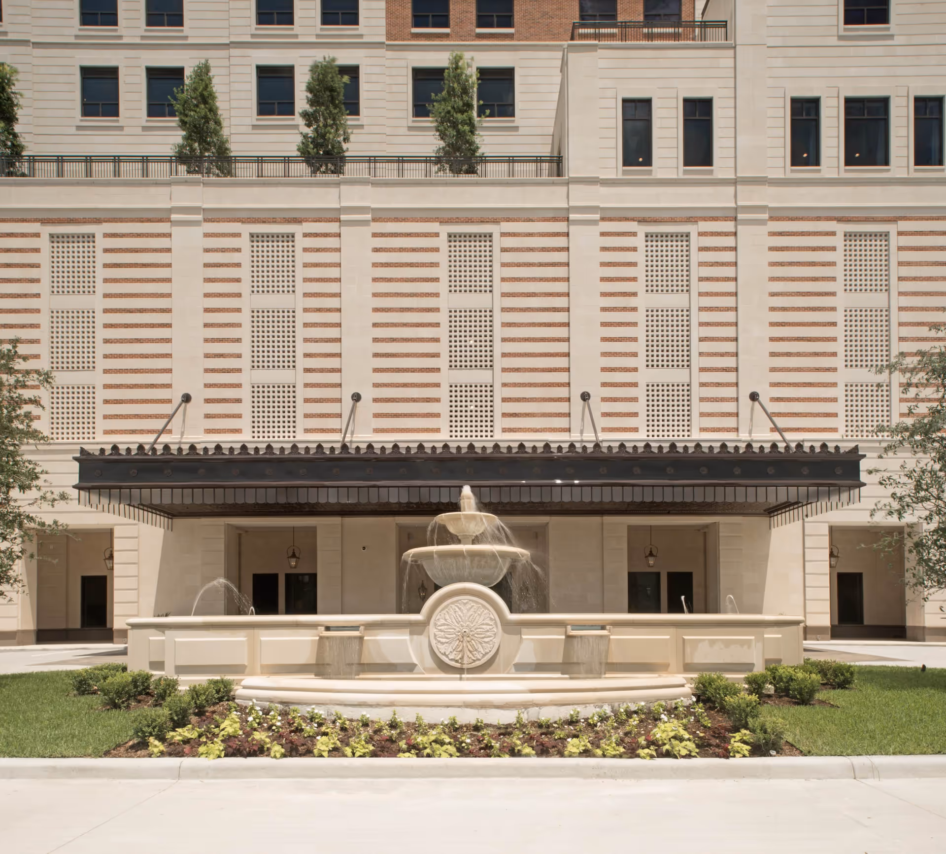 Front exterior view of a multi-story building with a decorative water fountain in the foreground, surrounded by landscaped greenery and flowers. The building features a beige facade with brick accents and several windows, along with a black awning above the entrance.