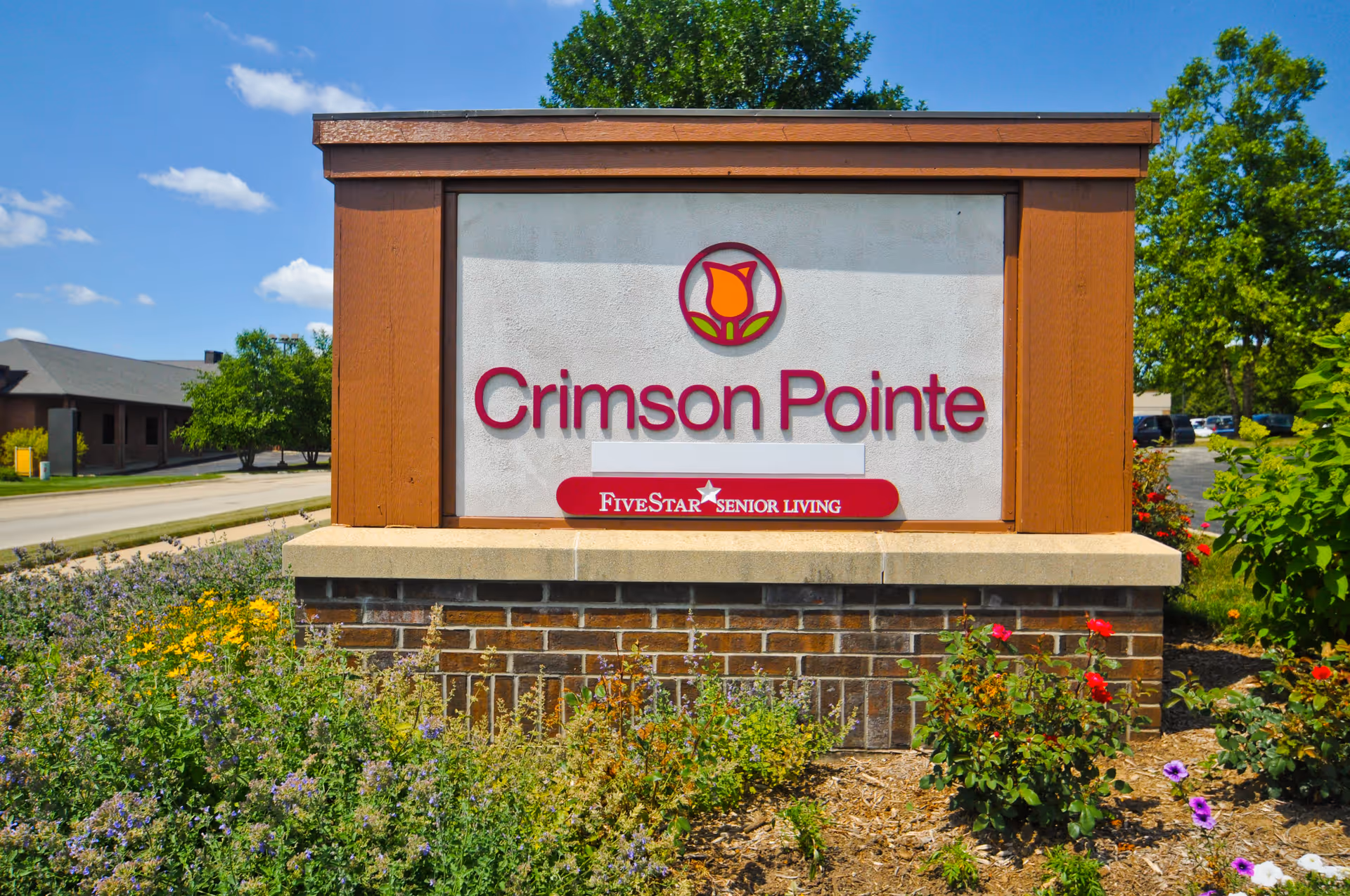 Outdoor sign for Crimson Pointe Five Star Senior Living with flowers and greenery in the foreground and a clear blue sky above.