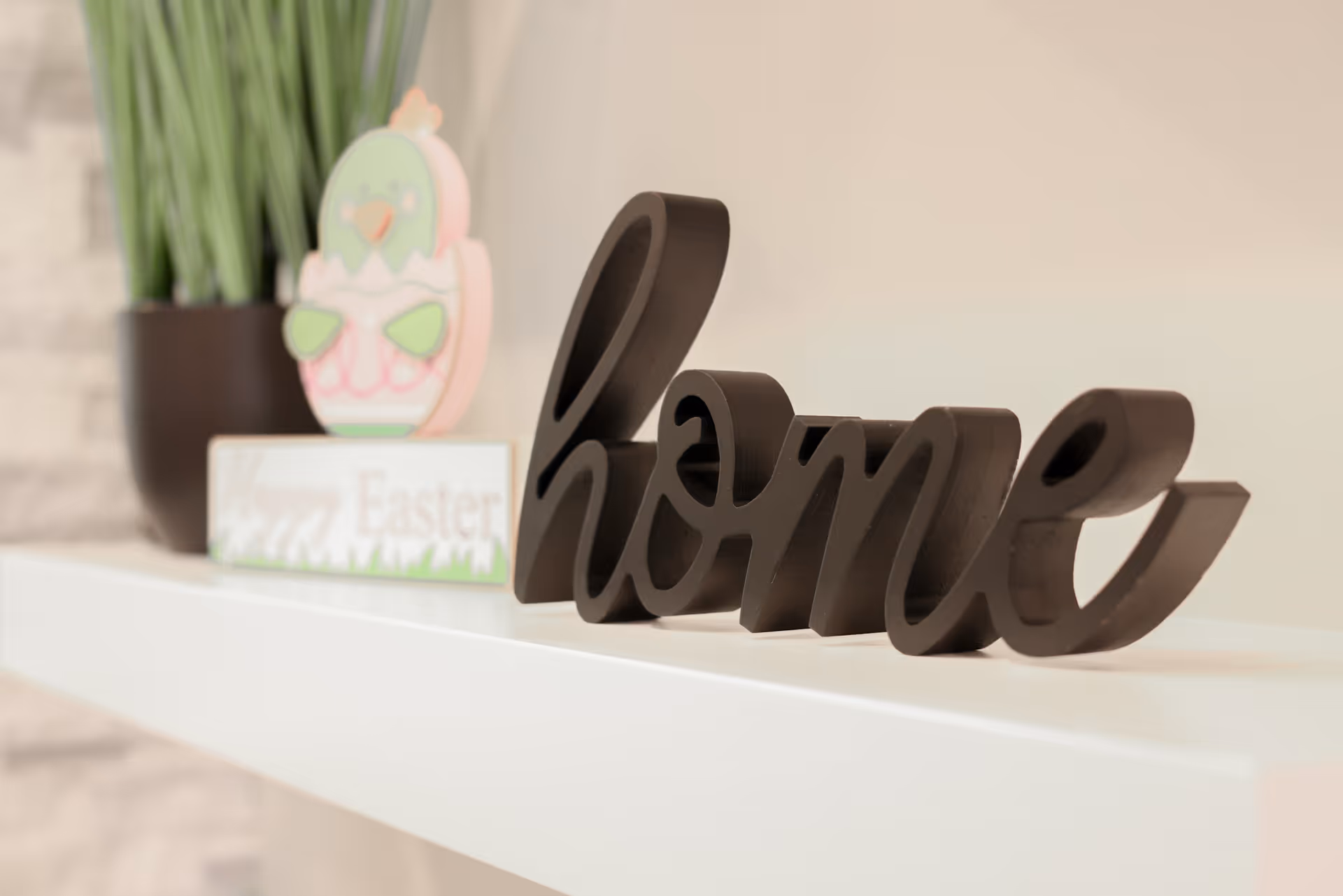 A white shelf displaying a black decorative 'home' sign with a potted plant and an Easter decoration behind it.