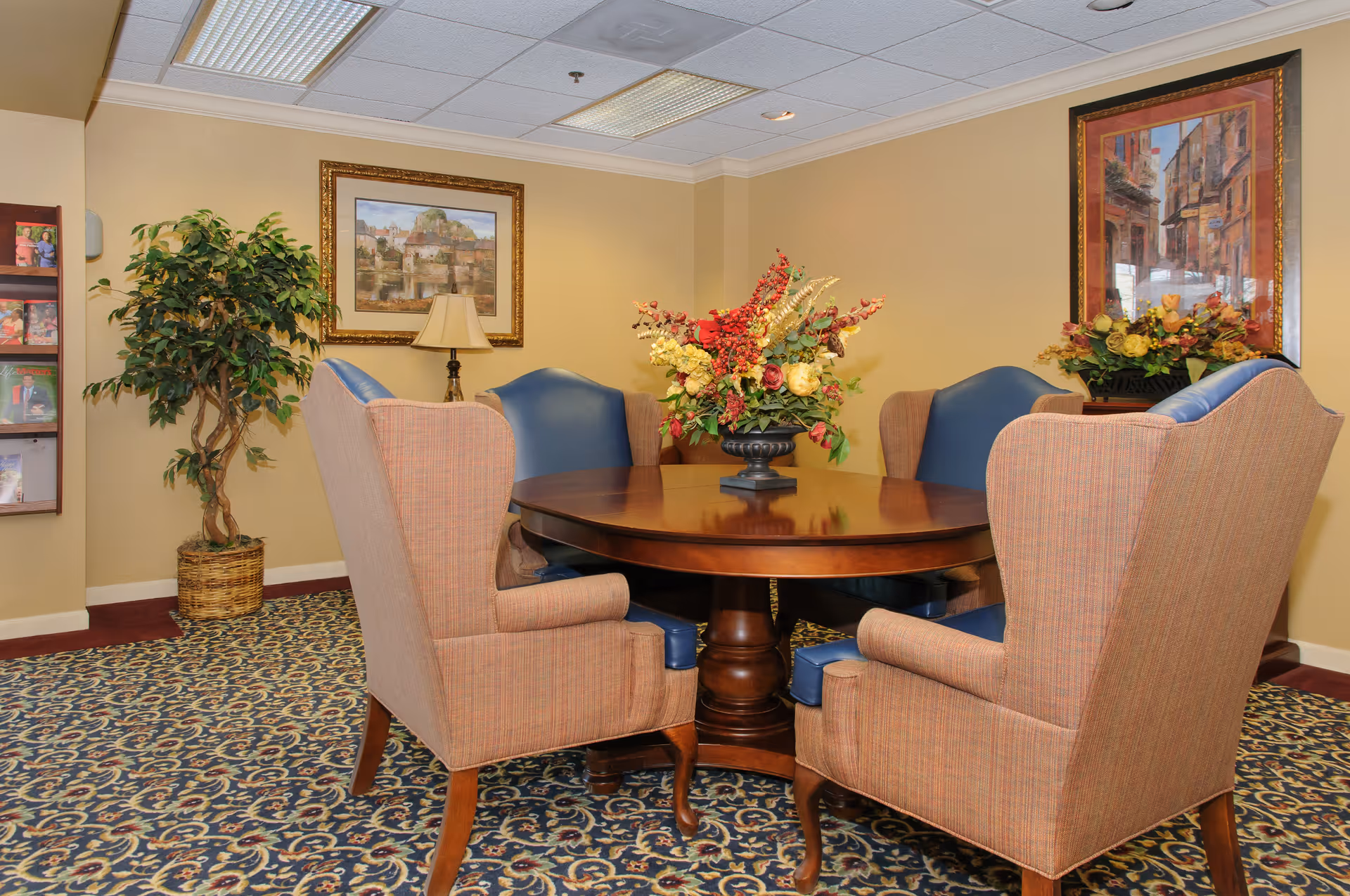 A cozy interior room with a round wooden table surrounded by six upholstered chairs, four with a beige fabric and two with blue leather. The table has a large floral centerpiece. The room features patterned carpet, beige walls, two framed paintings, a potted plant, a lamp, and a magazine rack on the wall.