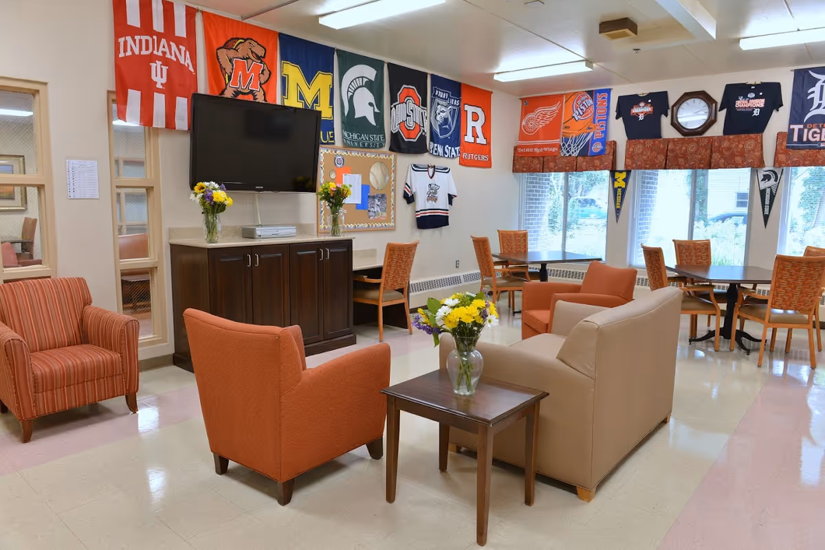 A bright and cozy common area with comfortable seating including orange and beige armchairs and a small wooden table with a vase of flowers. A flat-screen TV is mounted on the wall above a dark wooden cabinet. The walls are decorated with various college and sports team banners and jerseys. Large windows allow natural light to fill the room, and there are tables and chairs arranged near the windows.