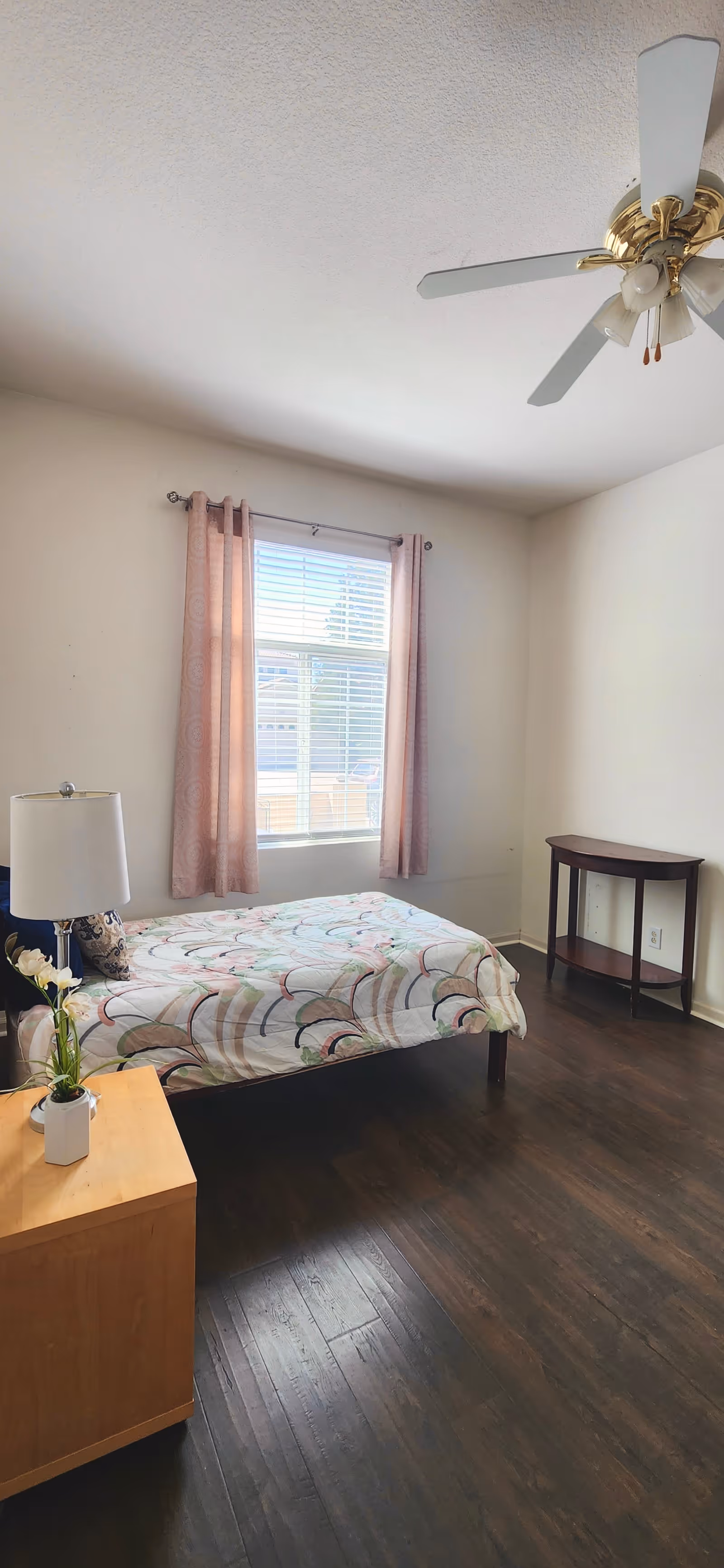 A simple bedroom with a single bed covered in a patterned bedspread, a wooden nightstand with a lamp and a small plant, a window with pink curtains, a ceiling fan with lights, and a small wooden table against the wall.