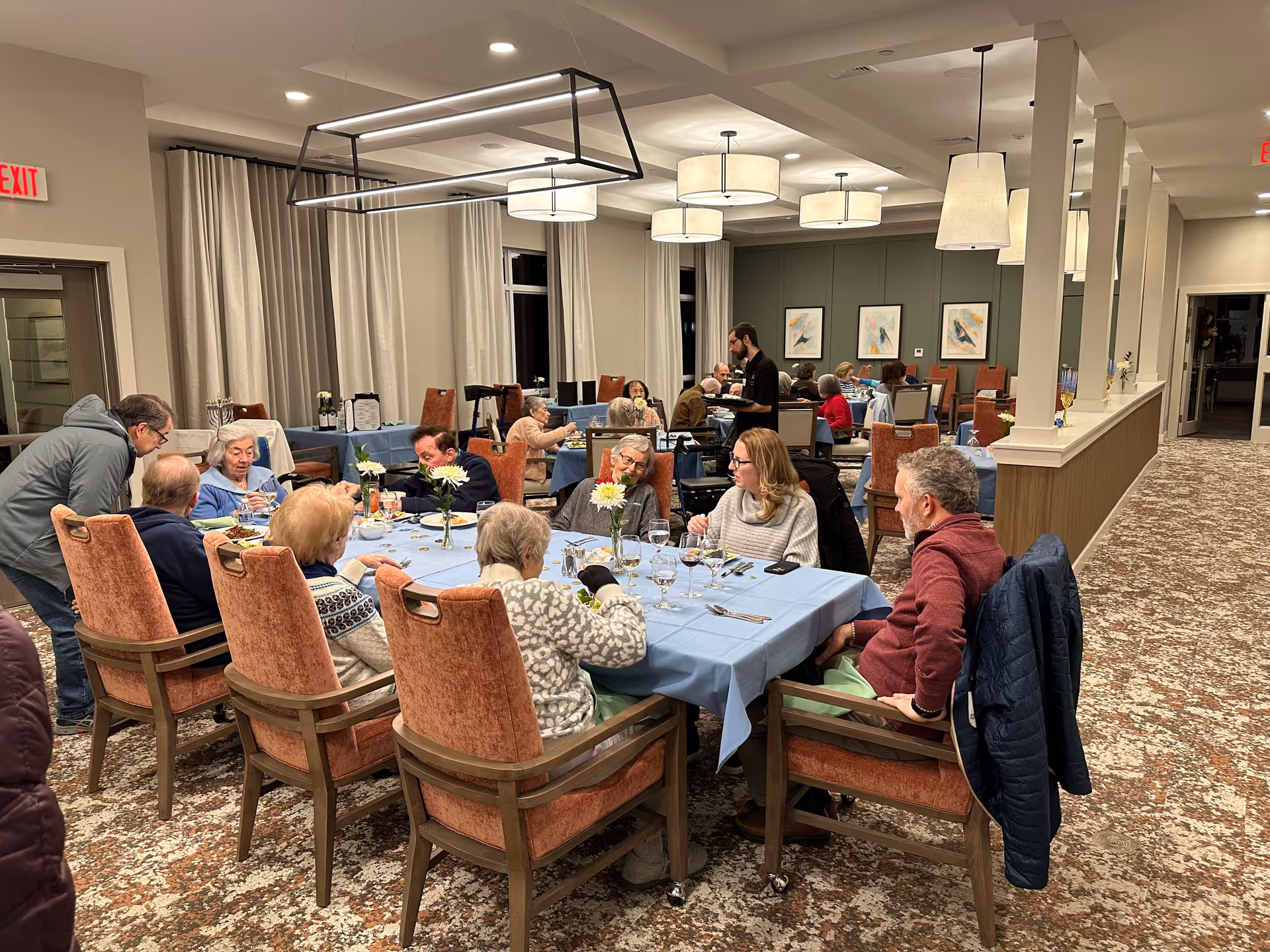A group of elderly people and a few younger adults sitting around tables with blue tablecloths in a dining room, enjoying a meal and conversation. The room has warm lighting with modern hanging light fixtures, beige curtains, and framed artwork on the walls. A staff member is serving food to the guests.