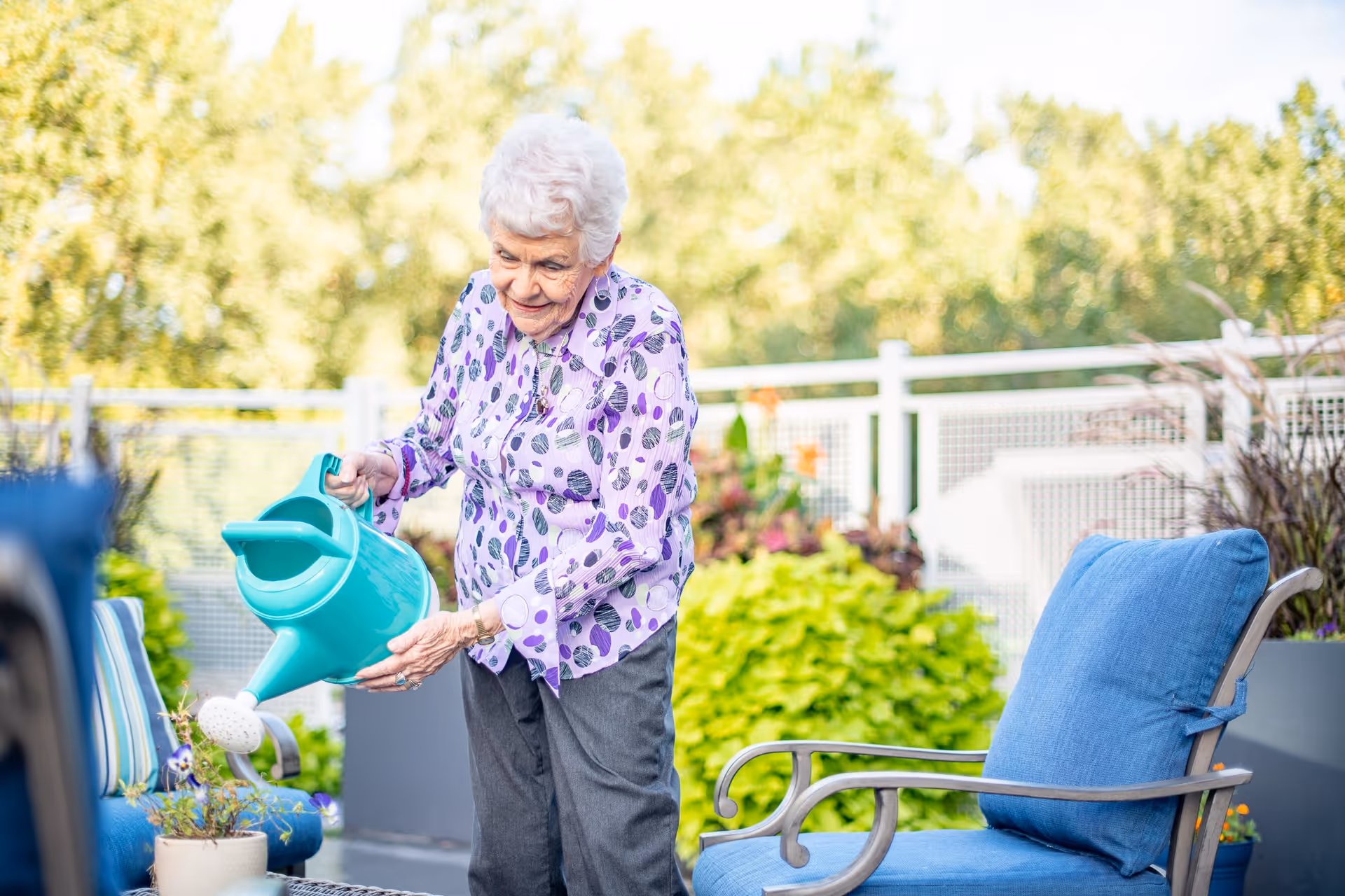 An elderly woman with white hair wearing a purple patterned blouse and gray pants is watering plants with a turquoise watering can on a patio. The patio has blue cushioned chairs and is surrounded by greenery and a white fence.