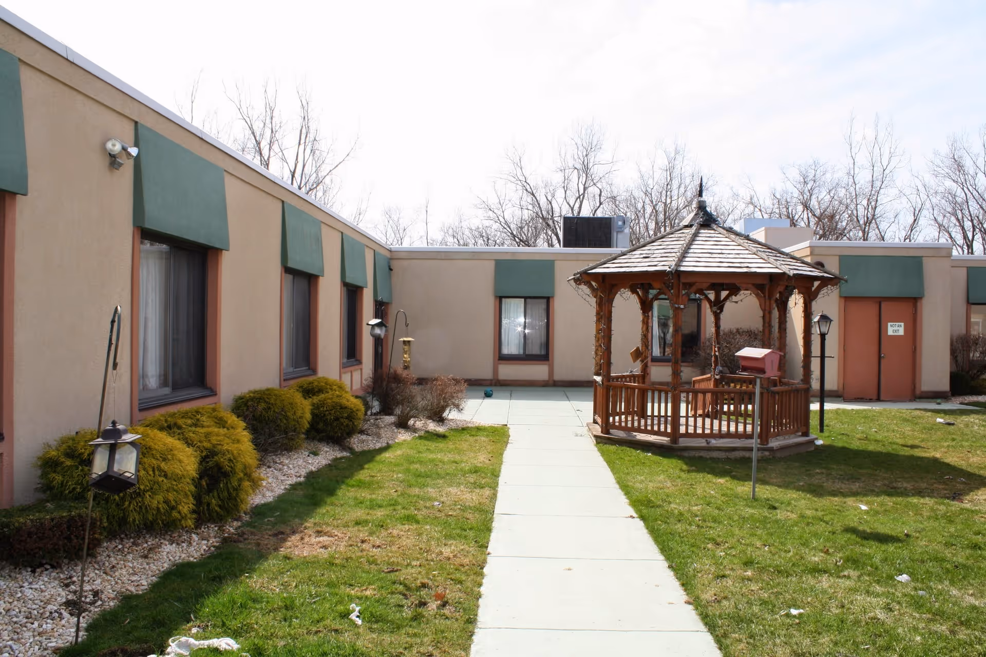 Outdoor courtyard area of a senior living facility with a paved walkway leading to a wooden gazebo. The building has beige walls with green awnings over the windows. There are bushes and grass surrounding the walkway and gazebo, with leafless trees in the background under a partly cloudy sky.