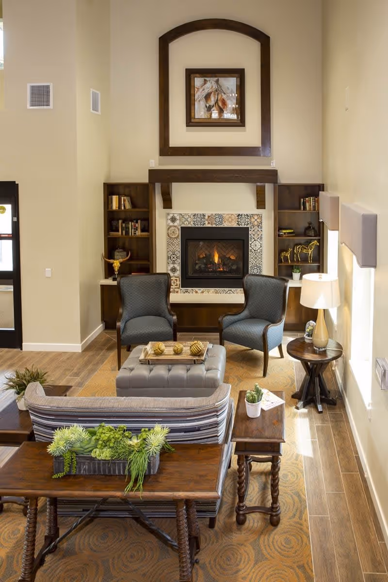 A cozy living room area with a fireplace surrounded by decorative tiles. Two blue upholstered armchairs face a tufted ottoman with decorative golden balls on a tray. Behind the ottoman is a striped sofa with a wooden console table holding a planter with green succulents. There are built-in wooden shelves on either side of the fireplace with books and decorative items. A side table with a lamp and a small plant is next to one of the armchairs. The room has wooden flooring and a patterned area rug.